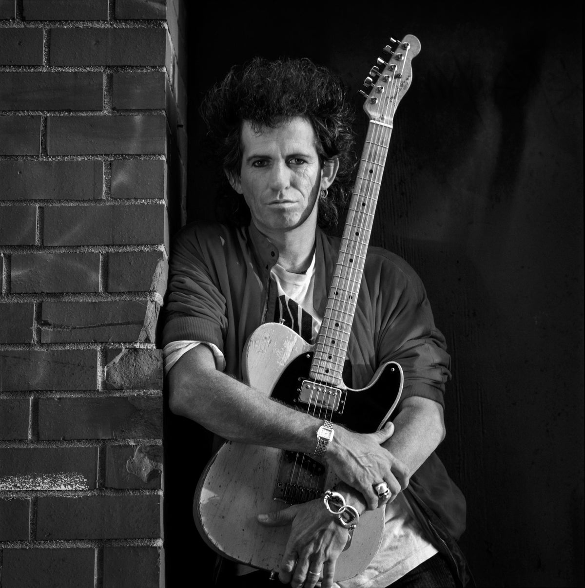 Keith Richards sits in a brick doorway holding an electric guitar upright against his body. Photograph by Timothy White, New York City, 1988.