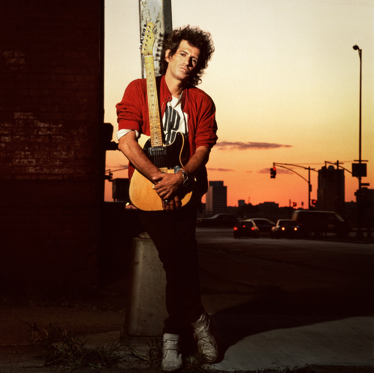 Keith Richards holds a guitar on a city street as the sky behind him turns warm with evening light. Photograph by Timothy White, New York City, 1988.