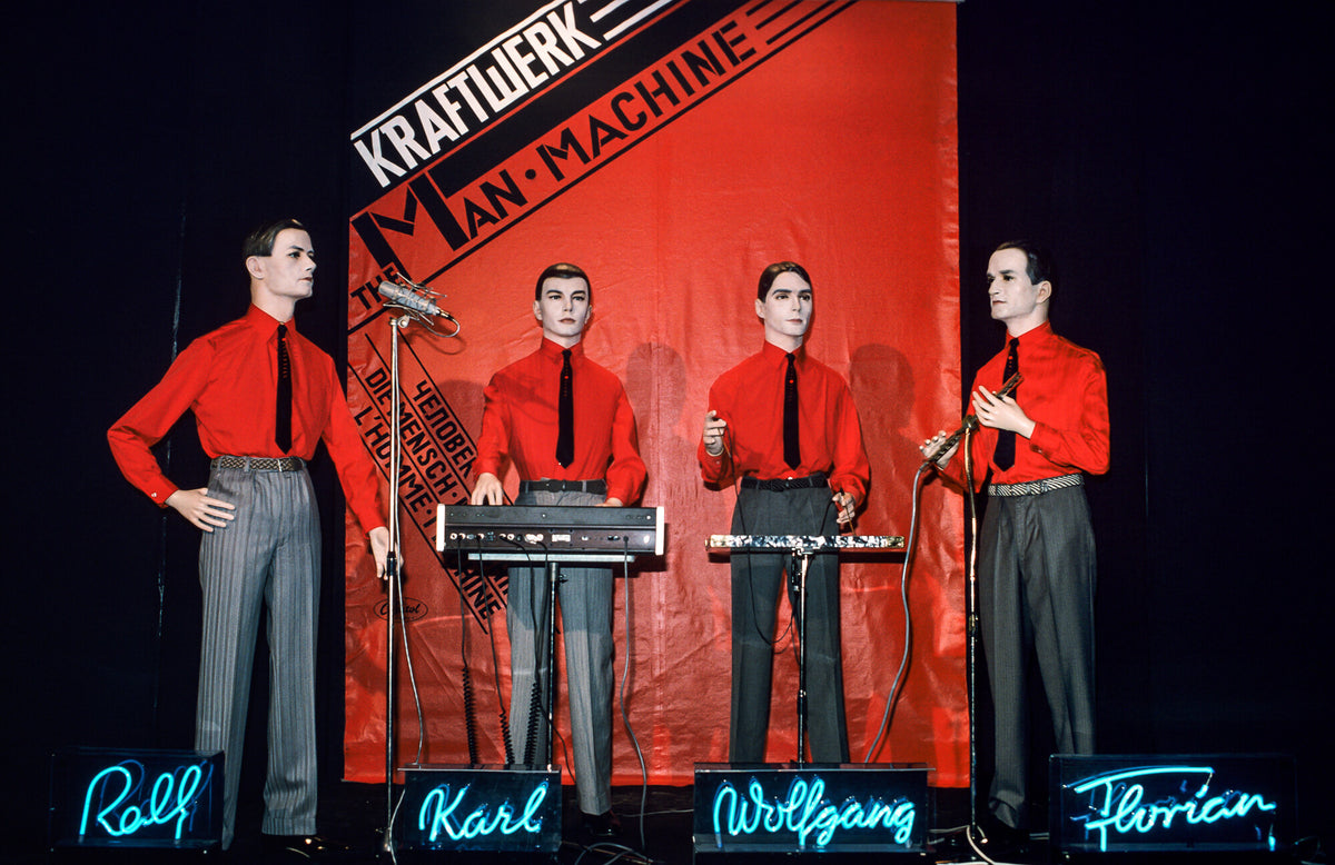 Kraftwerk figures in red shirts behind keyboards with blue neon names, 1978, photographed by Lynn Goldsmith.