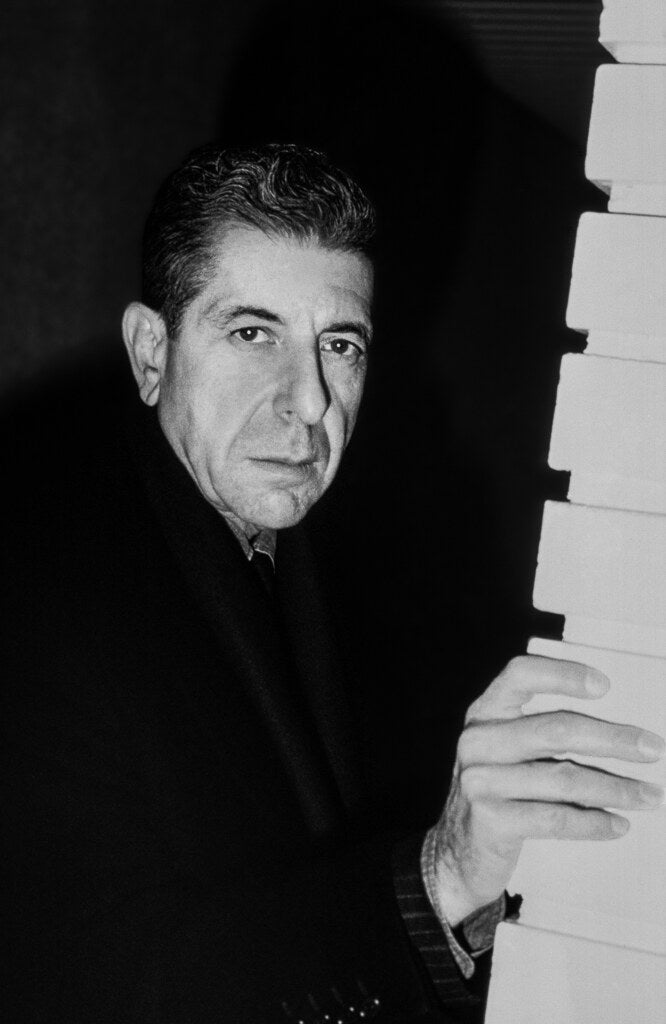 Leonard Cohen in an overcoat beside a piano keyboard, in black and white, 1988, photographed by Lynn Goldsmith.