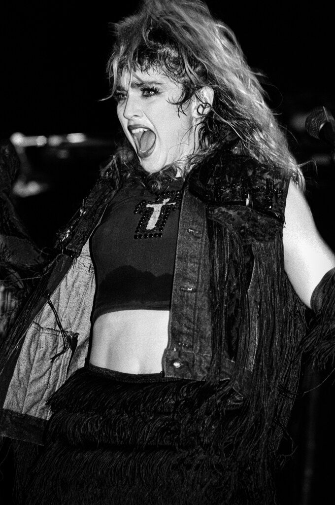 Madonna singing in a fringed jacket and crucifix, in black and white, 1985, photographed by Lynn Goldsmith.