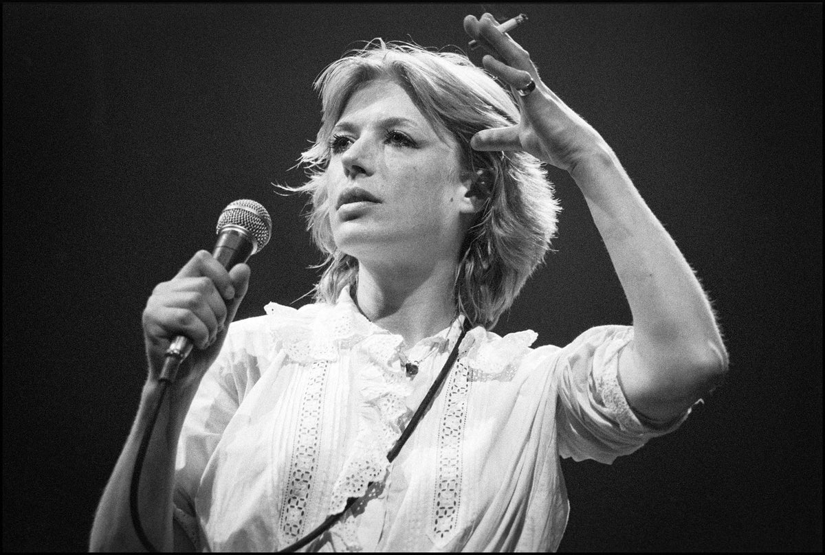 Marianne Faithfull singing into a microphone with a cigarette in her raised hand, wearing a white lace blouse, Dominion Theatre, London, 1982, photographed by David Corio.