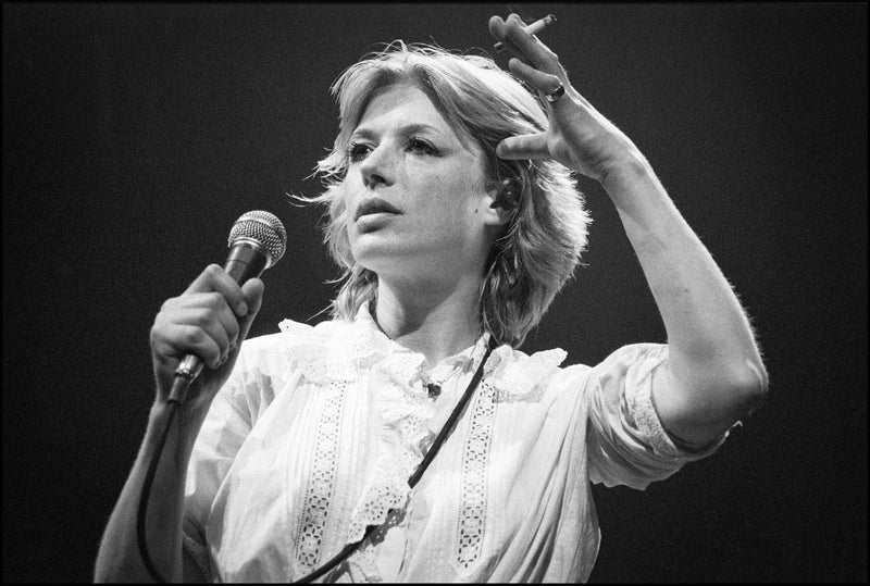 Marianne Faithfull singing into a microphone with a cigarette in her raised hand, wearing a white lace blouse, Dominion Theatre, London, 1982, photographed by David Corio.