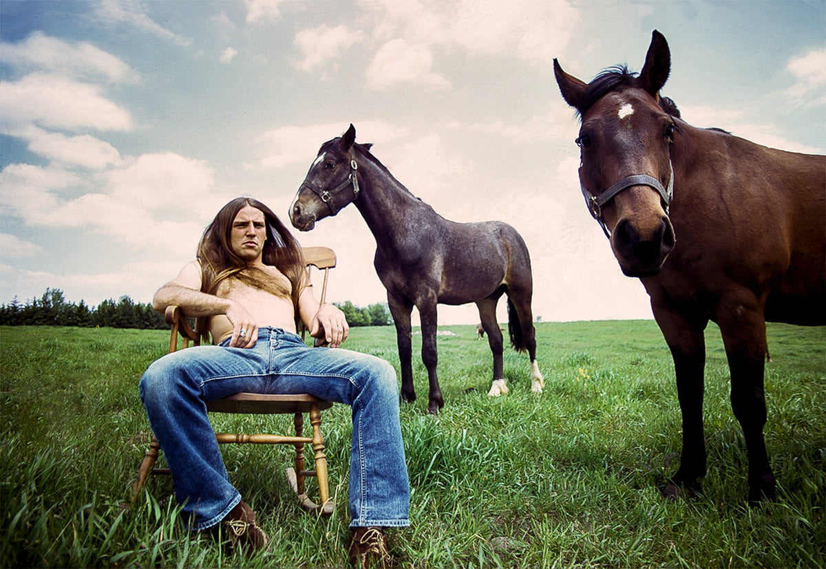 Mark Farner of Grand Funk Railroad seated in a field beside two horses, in color, 1973, photographed by Lynn Goldsmith.