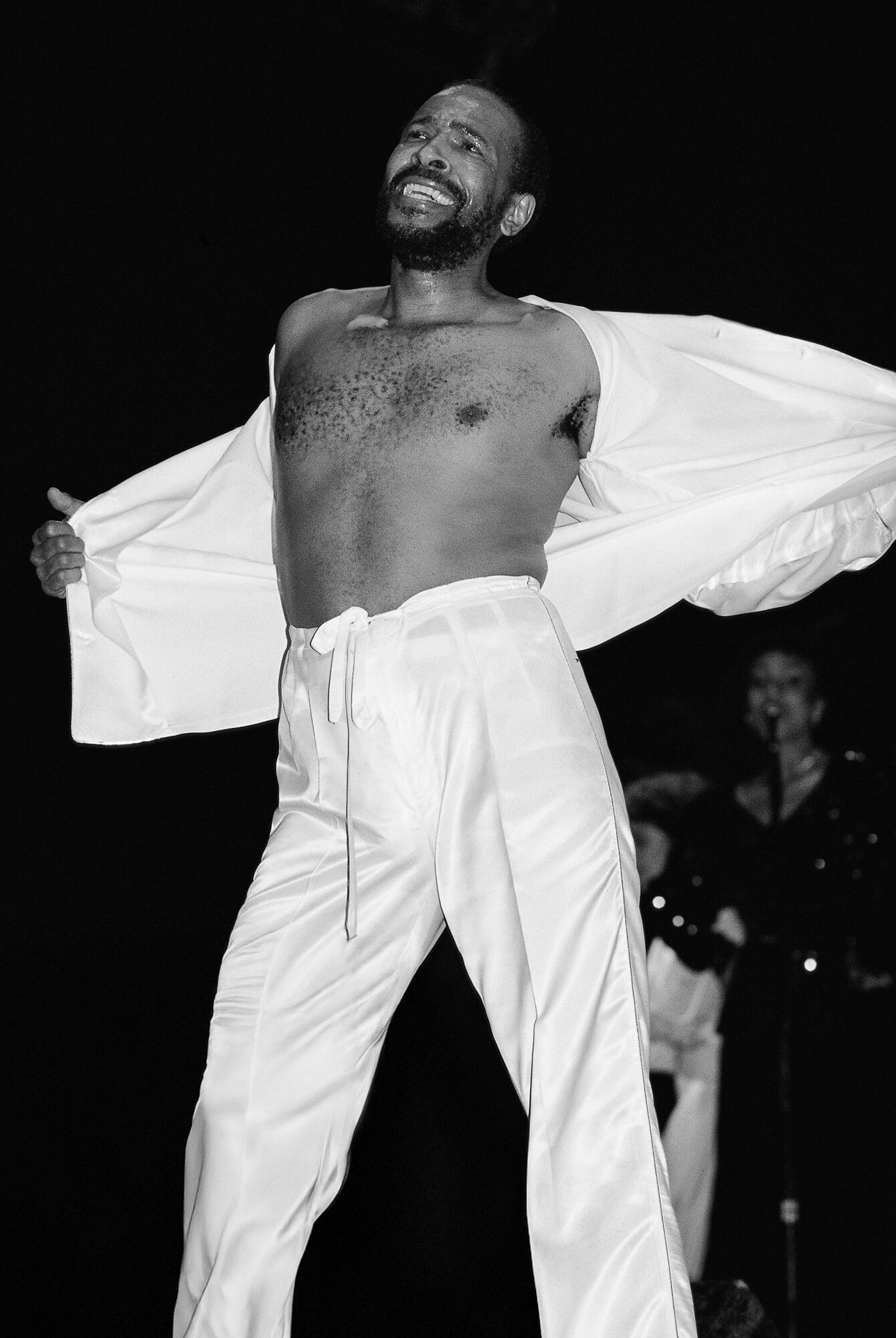 Marvin Gaye on stage in white, arms spread wide, in black and white, 1983, photographed by Lynn Goldsmith.