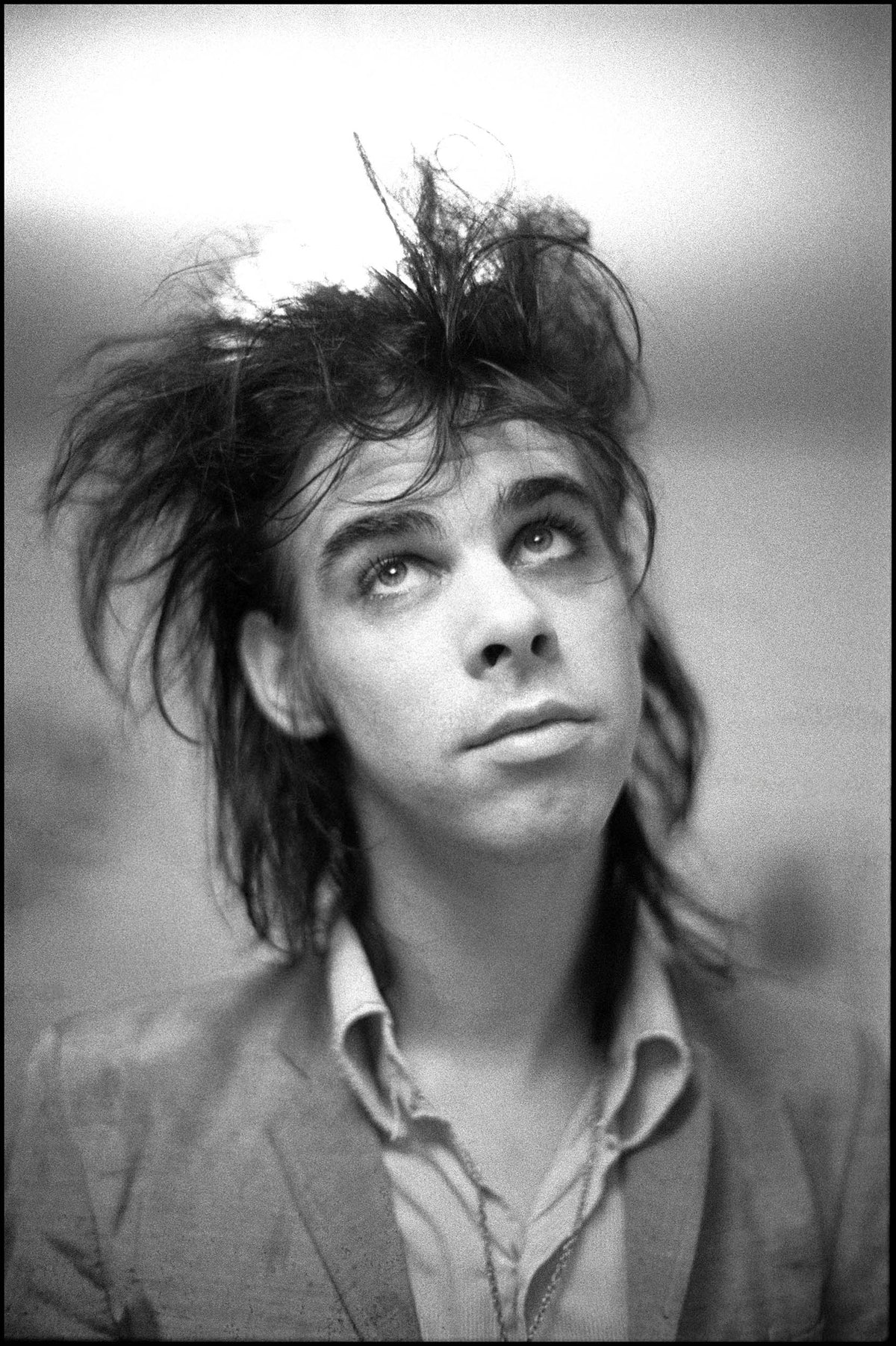 Close-up portrait of a young Nick Cave with wild hair looking upward, Kilburn, London, 1982, photographed by David Corio.