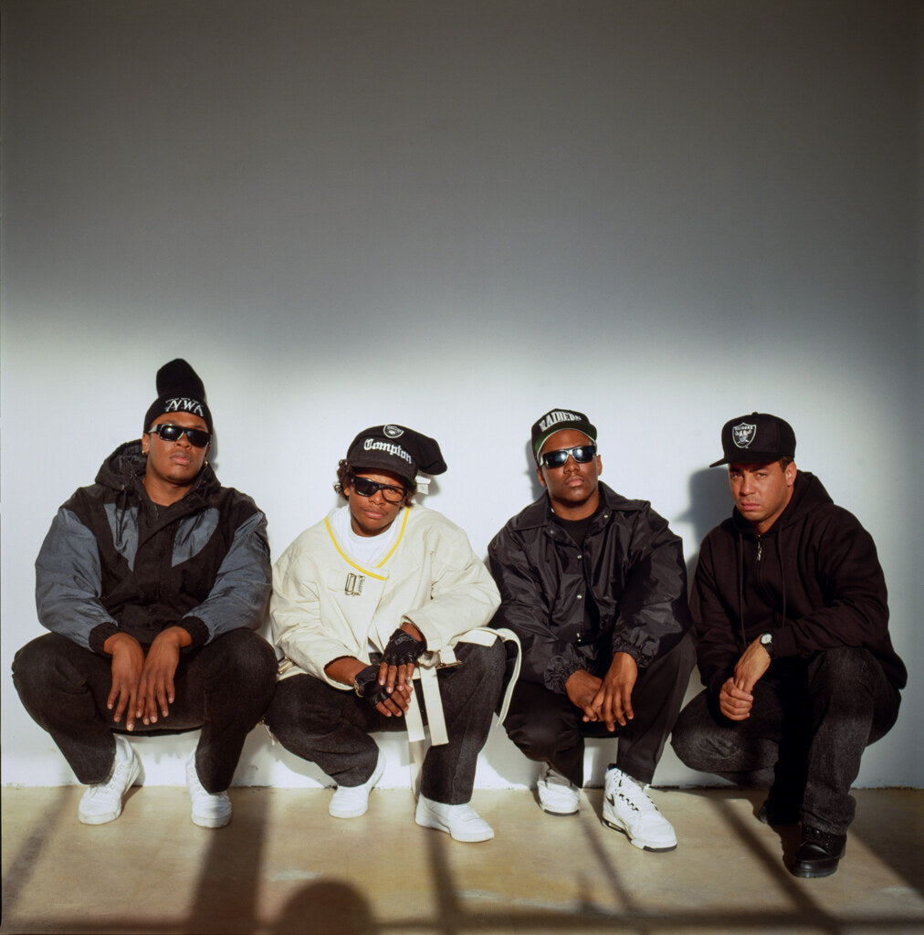 N.W.A crouched side by side against a white wall, in color, 1990, photographed by Lynn Goldsmith.