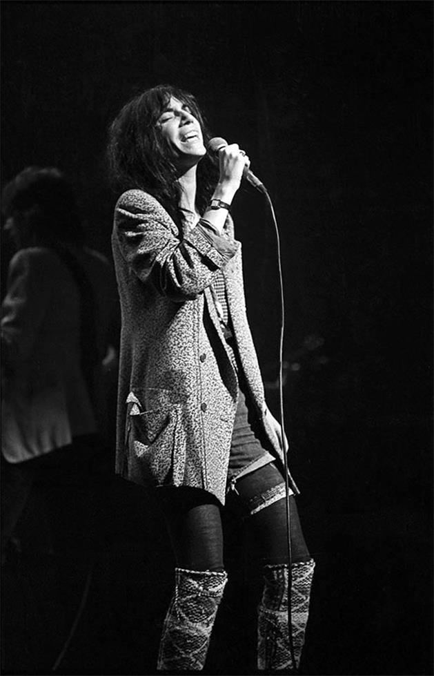 Patti Smith on stage at the microphone in a patterned coat, photographed by Charlyn Zlotnik, 1977.