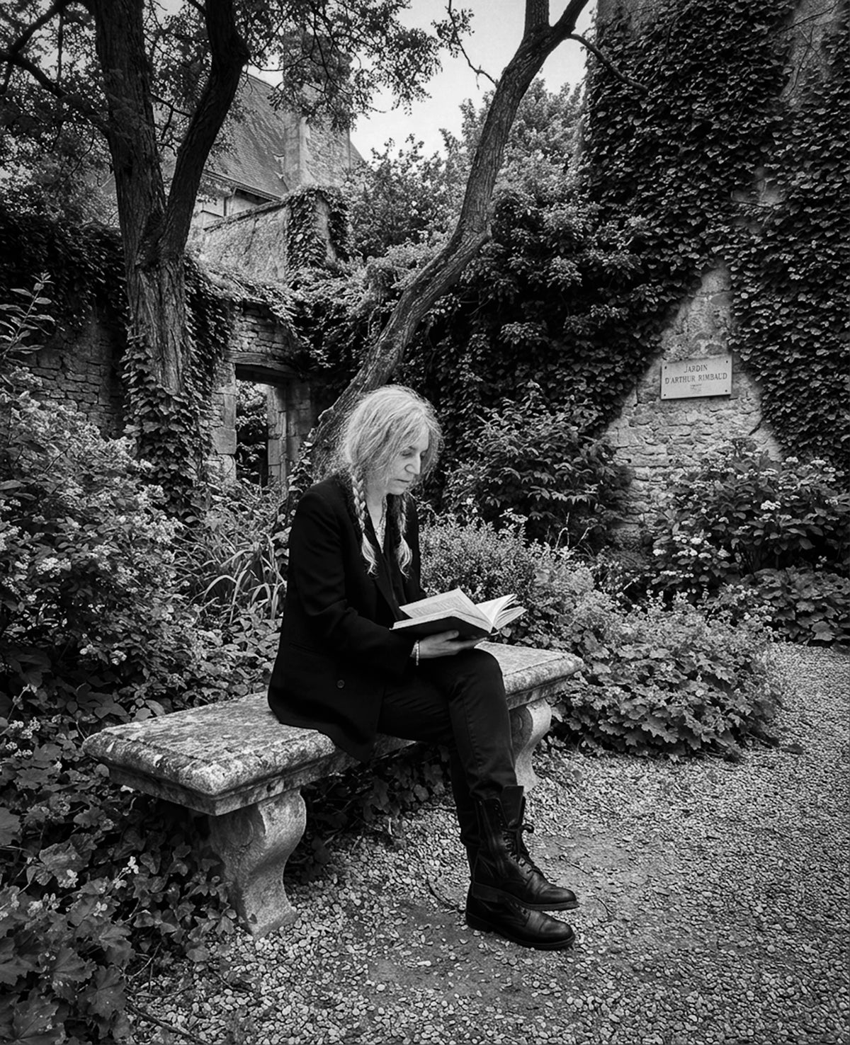 Patti Smith reading a book on a stone bench in a walled garden, photographed by Lynn Goldsmith.