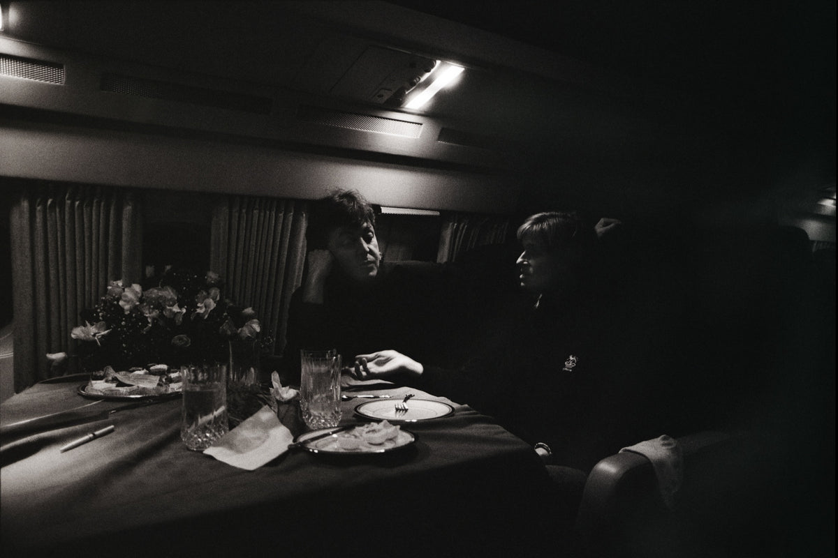Paul and Linda McCartney share a table inside an aircraft cabin in a low-lit, informal scene. Photograph by Timothy White, Toronto, Canada, 1989.