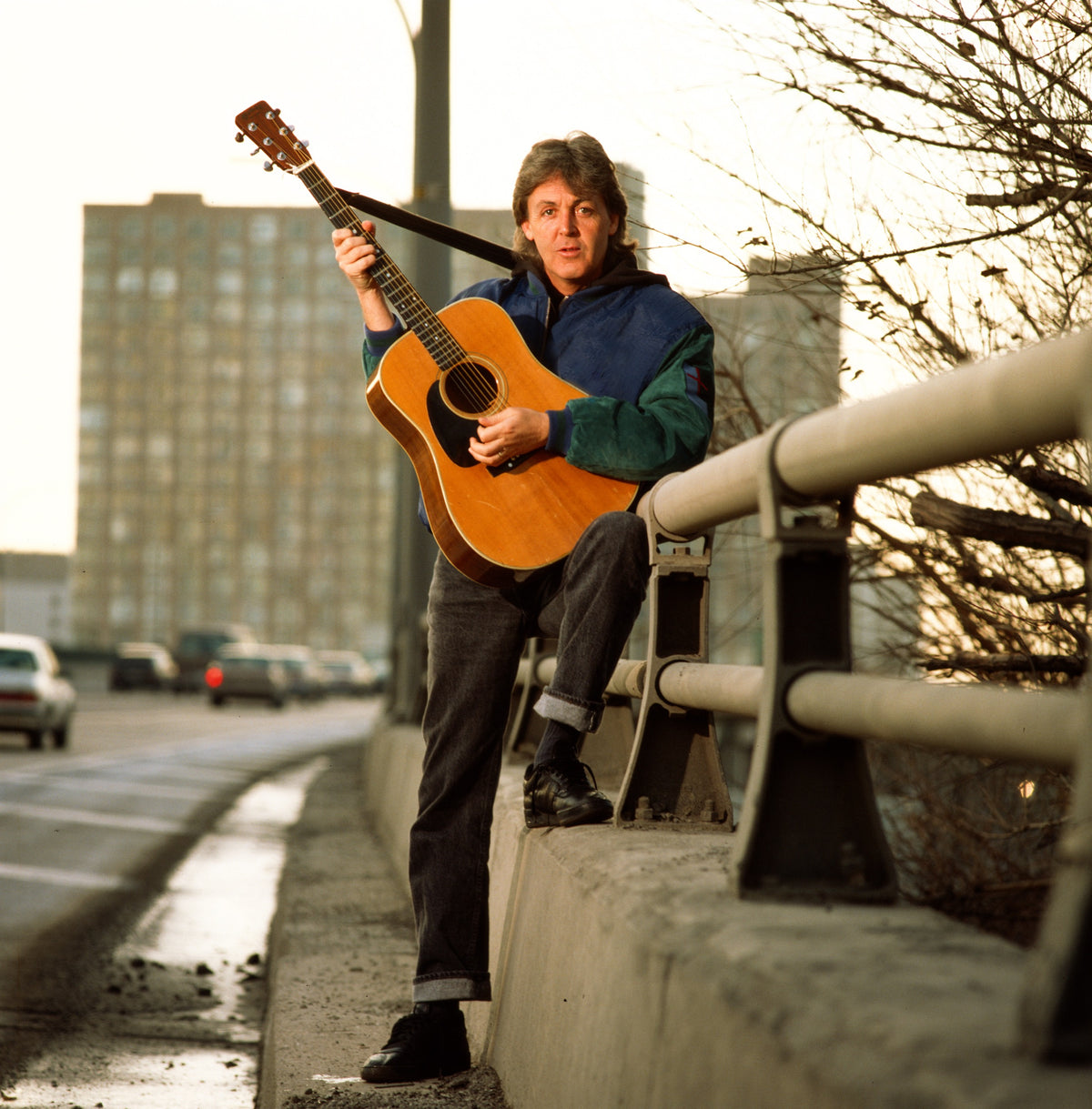 Paul McCartney holds an acoustic guitar beside a roadside railing, with the city behind him. Photograph by Timothy White, Toronto, Canada, 1989.