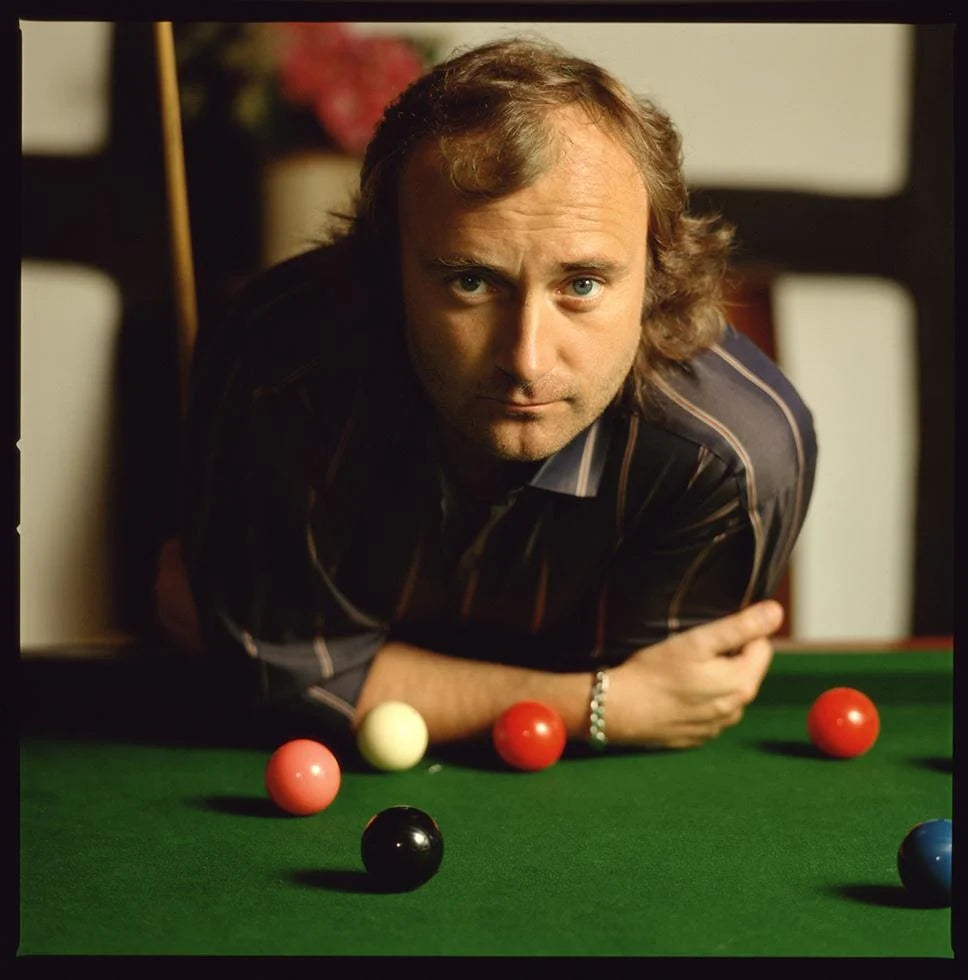 Phil Collins leaning over a green snooker table, photographed by Timothy White in London, England, 1986.