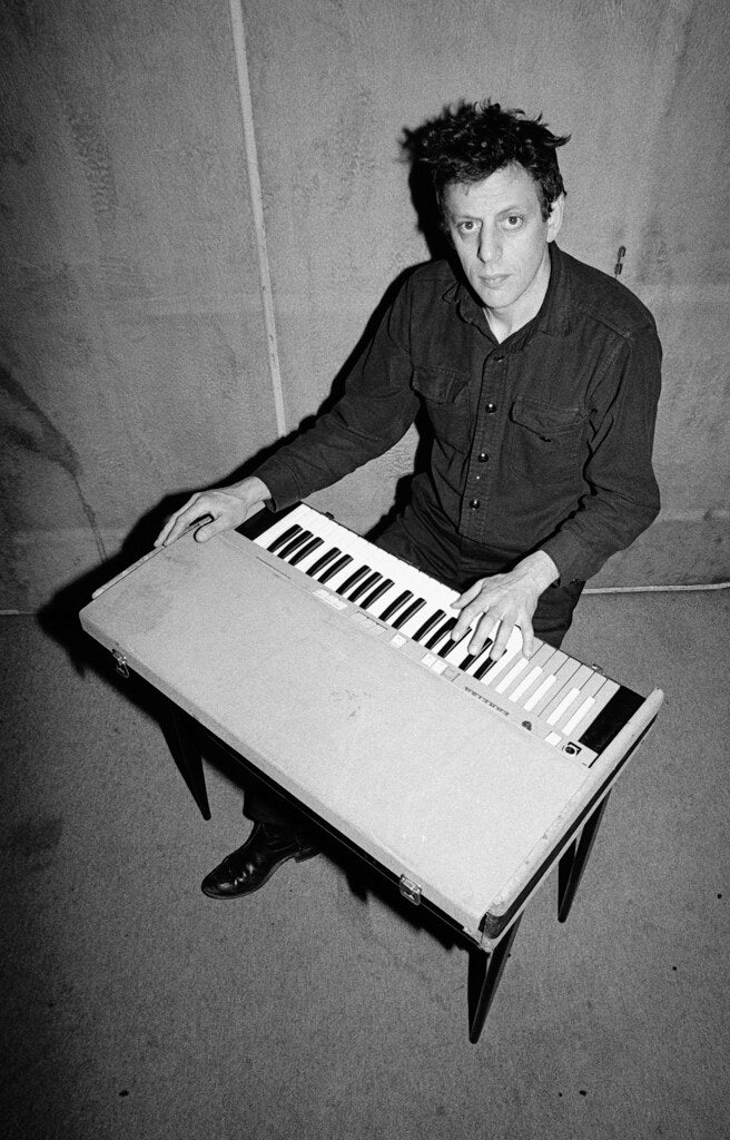 Philip Glass seated on the floor with a keyboard across his lap, in black and white, 1975, photographed by Lynn Goldsmith.
