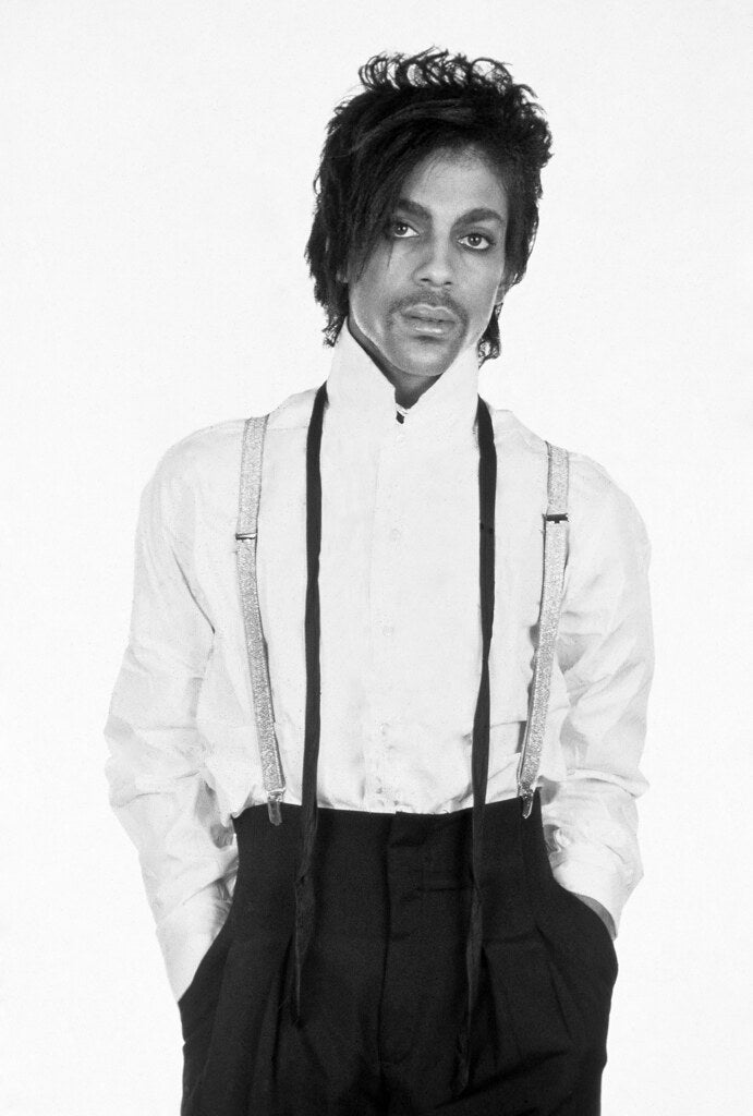 Prince in a white shirt and suspenders against a white backdrop, in black and white, 1981, photographed by Lynn Goldsmith.