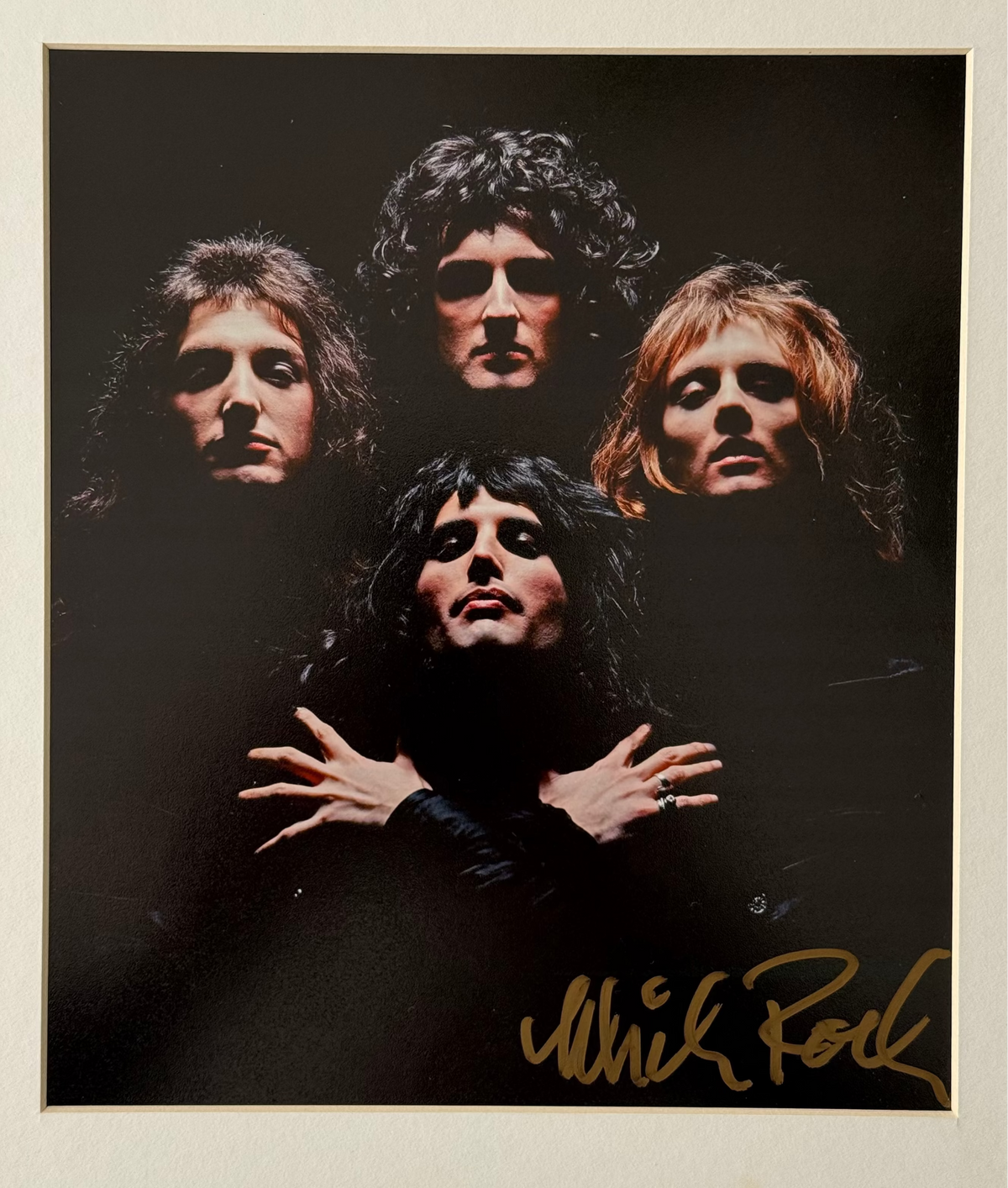 A signed photo print of Queen by Mick Rock, featuring four band members against a dark background with a gold signature in the bottom right corner, mounted on a white mat board.