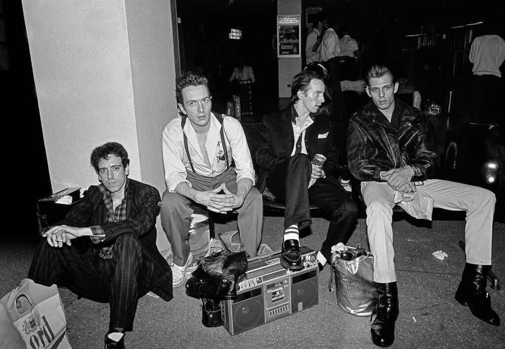 The Clash seated on a sidewalk with a boombox, in black and white, 1981, photographed by Lynn Goldsmith.