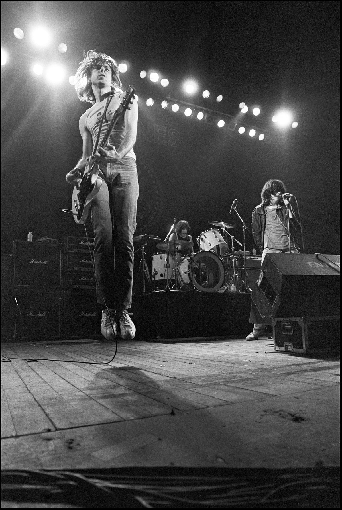 The Ramones performing under a row of stage lights with Johnny Ramone jumping in the air, Hammersmith Odeon, London, 1980, photographed by David Corio.