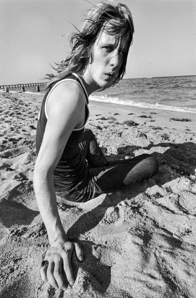 Todd Rundgren reclining on the sand with a pier behind him, in black and white, 1972, photographed by Lynn Goldsmith.