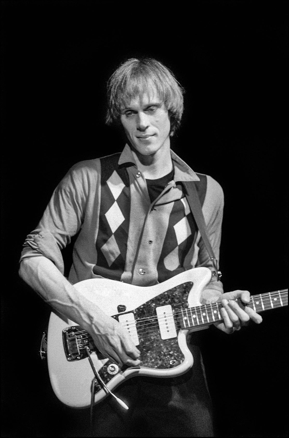 Tom Verlaine playing a Fender Jazzmaster guitar in a diamond-patterned vest on stage at The Venue, London, 1982, photographed by David Corio.