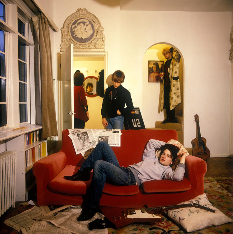 A young U2 lounging in a Bayswater flat, one member stretched across a red sofa with the band's name on a flight case, photographed by Martyn Goddard, London, 1979.