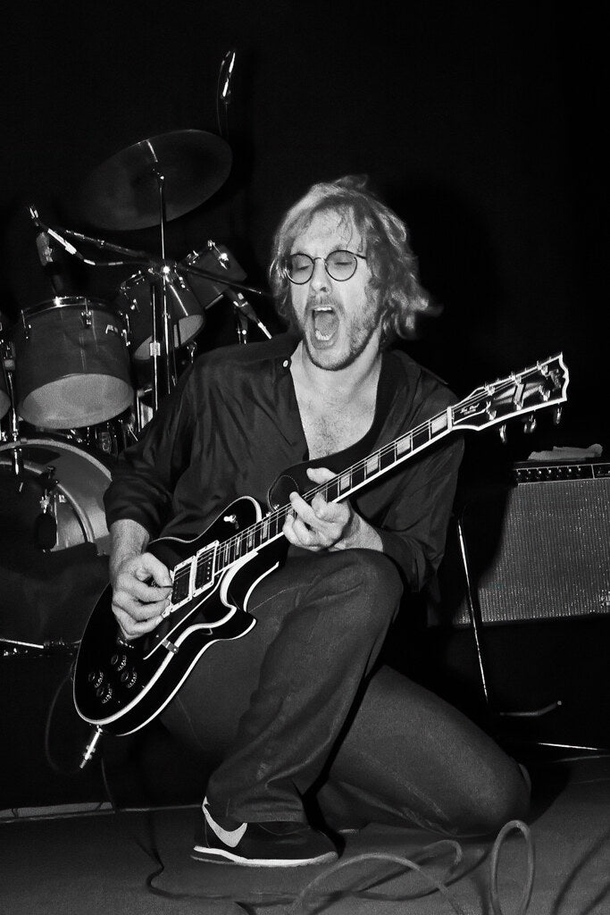 Warren Zevon crouching with his guitar on stage, in black and white, 1982, photographed by Lynn Goldsmith.