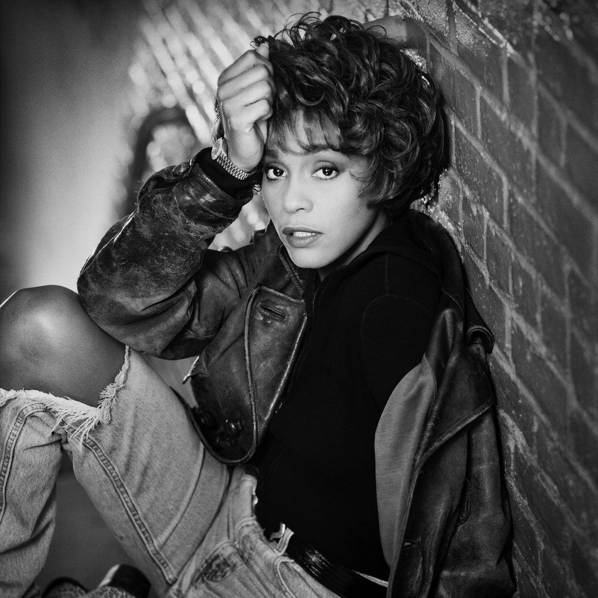 Whitney Houston sits against a brick wall, resting a hand against her forehead in this close portrait. Photograph by Timothy White, New York City, 1990.