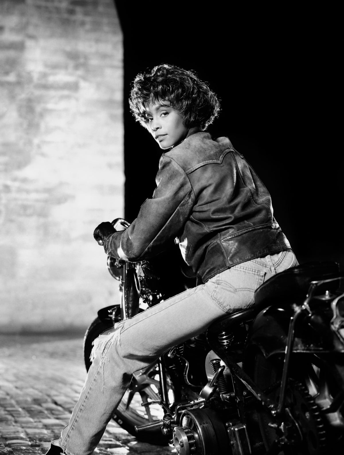 Whitney Houston sits on a motorcycle, turning back toward the camera. Photograph by Timothy White, Brooklyn, New York, 1990.