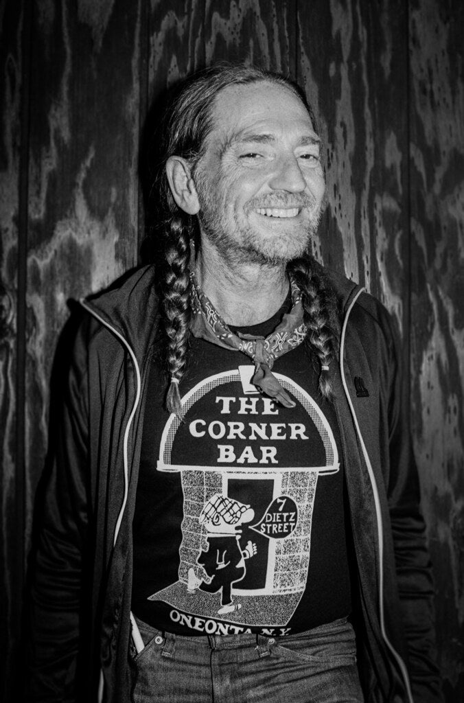 Willie Nelson smiling in braids and a Corner Bar T-shirt, in black and white, 1978, photographed by Lynn Goldsmith.