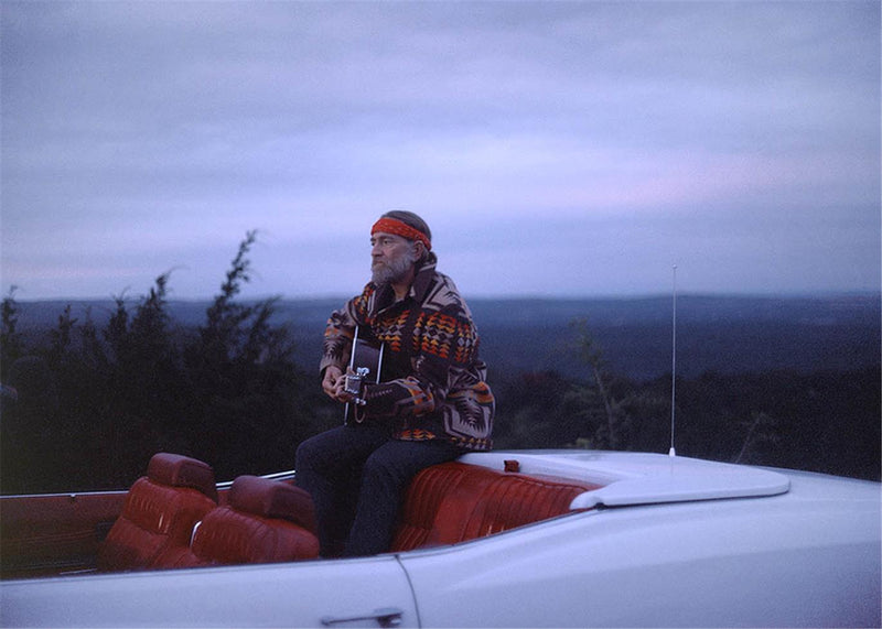 Willie Nelson playing guitar at sunset on a white convertible outside Austin, Texas, photographed by Charlyn Zlotnik, 1983.