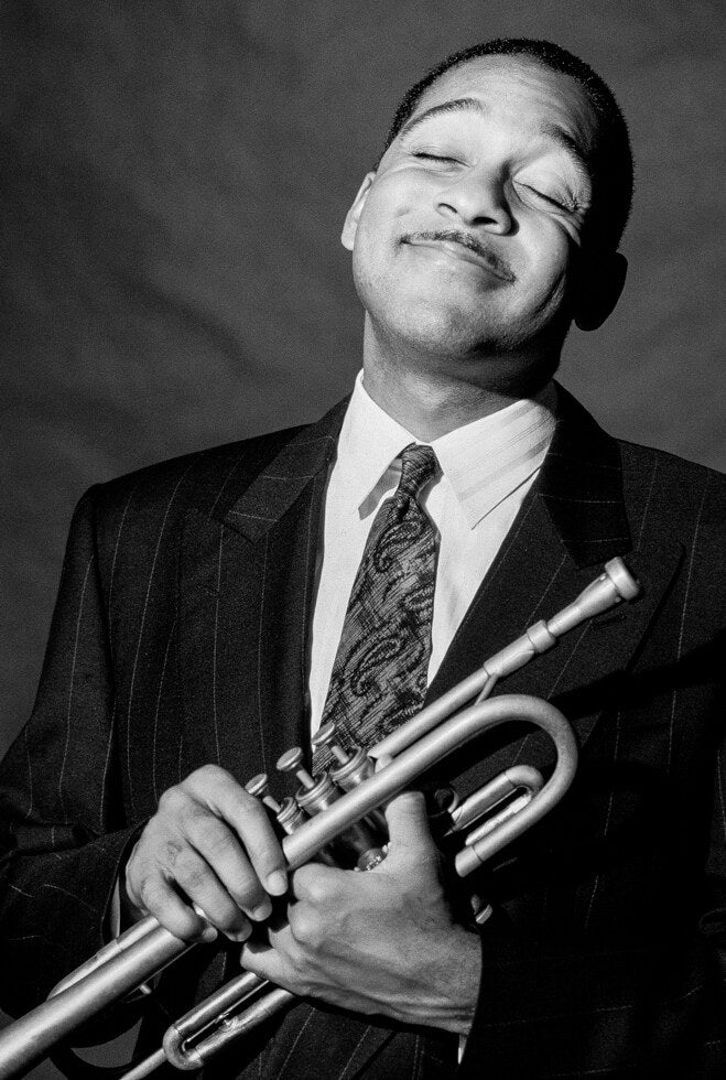 Wynton Marsalis smiling with his trumpet held to his chest, in black and white, 1990, photographed by Lynn Goldsmith.